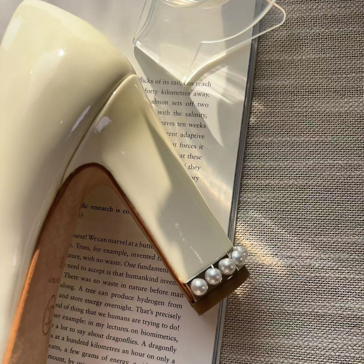 A close-up of a white patent leather clutch purse with a pearl-embellished clasp, resting on an open book over a gray textured fabric background.