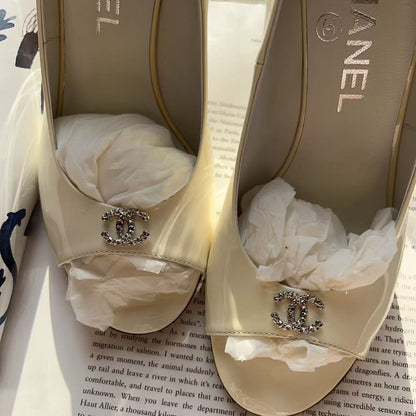 A pair of Chanel low-heel mule sandals in beige satin with crystal-embellished double-C logos, resting on top of a white Chanel shoebox and printed book pages.