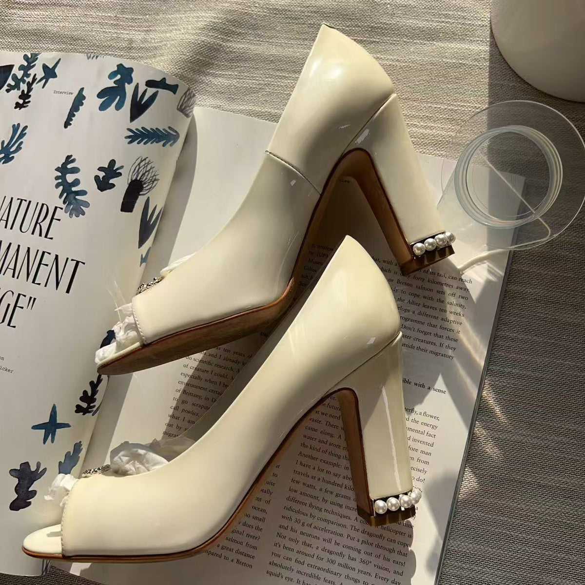 A pair of glossy off-white peep-toe high heels with block heels and pearl embellishments on the heels, styled on top of an open book with a glass and a ceramic cup nearby.