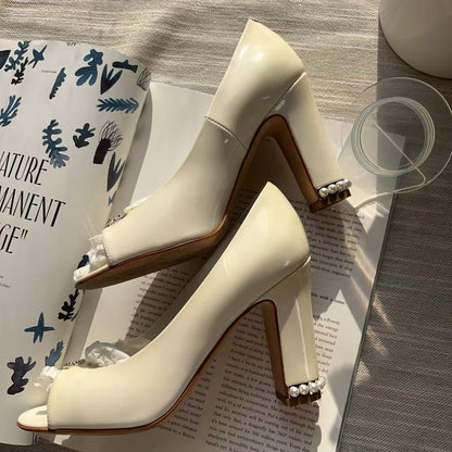 A pair of glossy off-white peep-toe high heels with block heels and pearl embellishments on the heels, styled on top of an open book with a glass and a ceramic cup nearby.