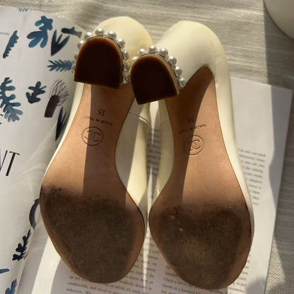 A pair of women's low-heeled mule shoes shown from the bottom, featuring a cream-colored leather upper, a brown insole, and a decorative border of white faux pearls around the heel strap. The shoes are styled on top of printed books on a gray textured surface.