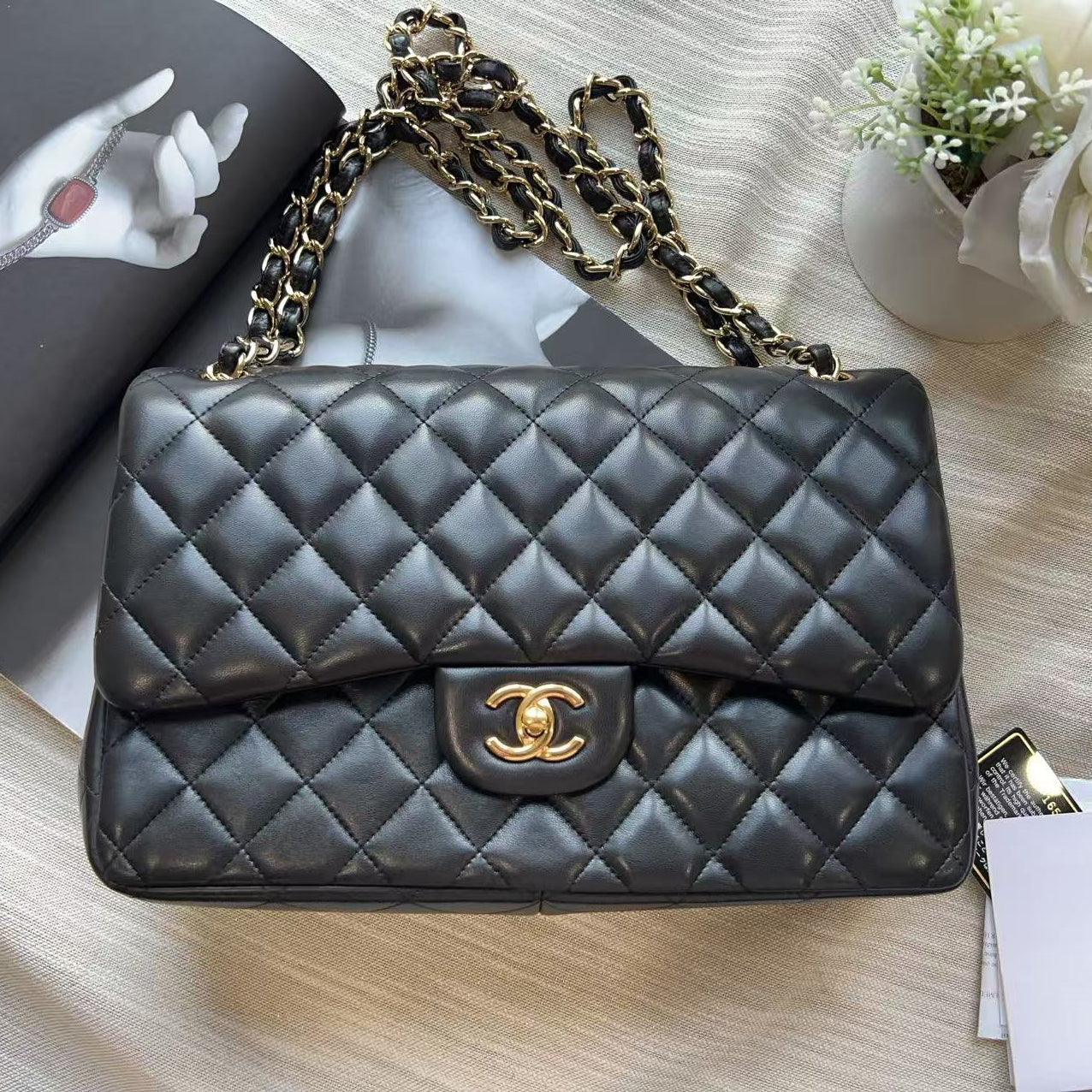 A black quilted Chanel shoulder bag with a gold-tone CC turn-lock closure and a gold chain strap, styled on a beige fabric surface with a magazine, white card, and white flower.