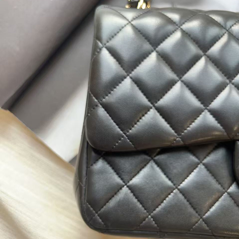 Close-up of a black quilted leather handbag with a gold chain strap, resting on a light wood surface next to a gray box.
