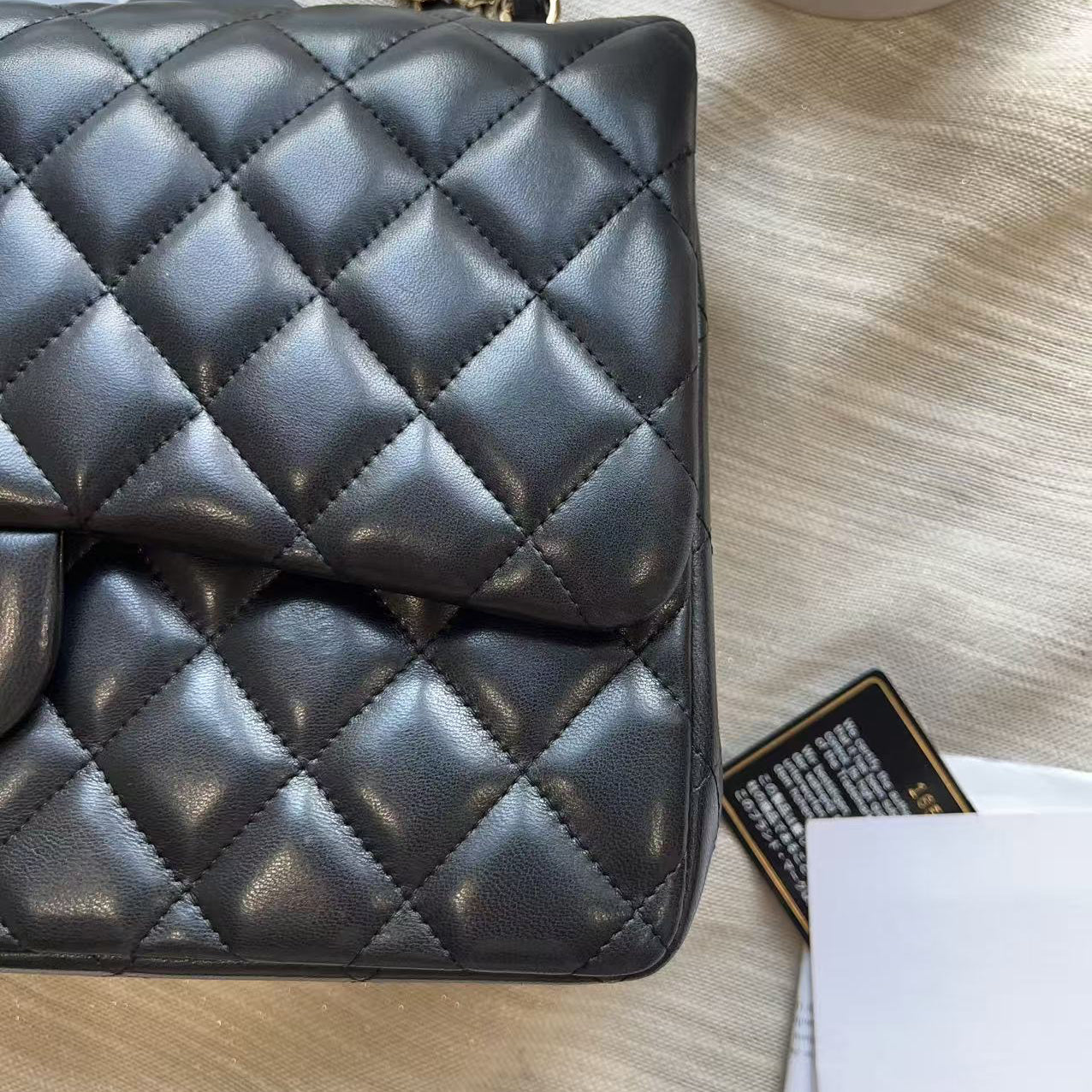 A close-up of a black quilted leather handbag with a gold chain strap, placed on a light wood surface with a tag and papers nearby.