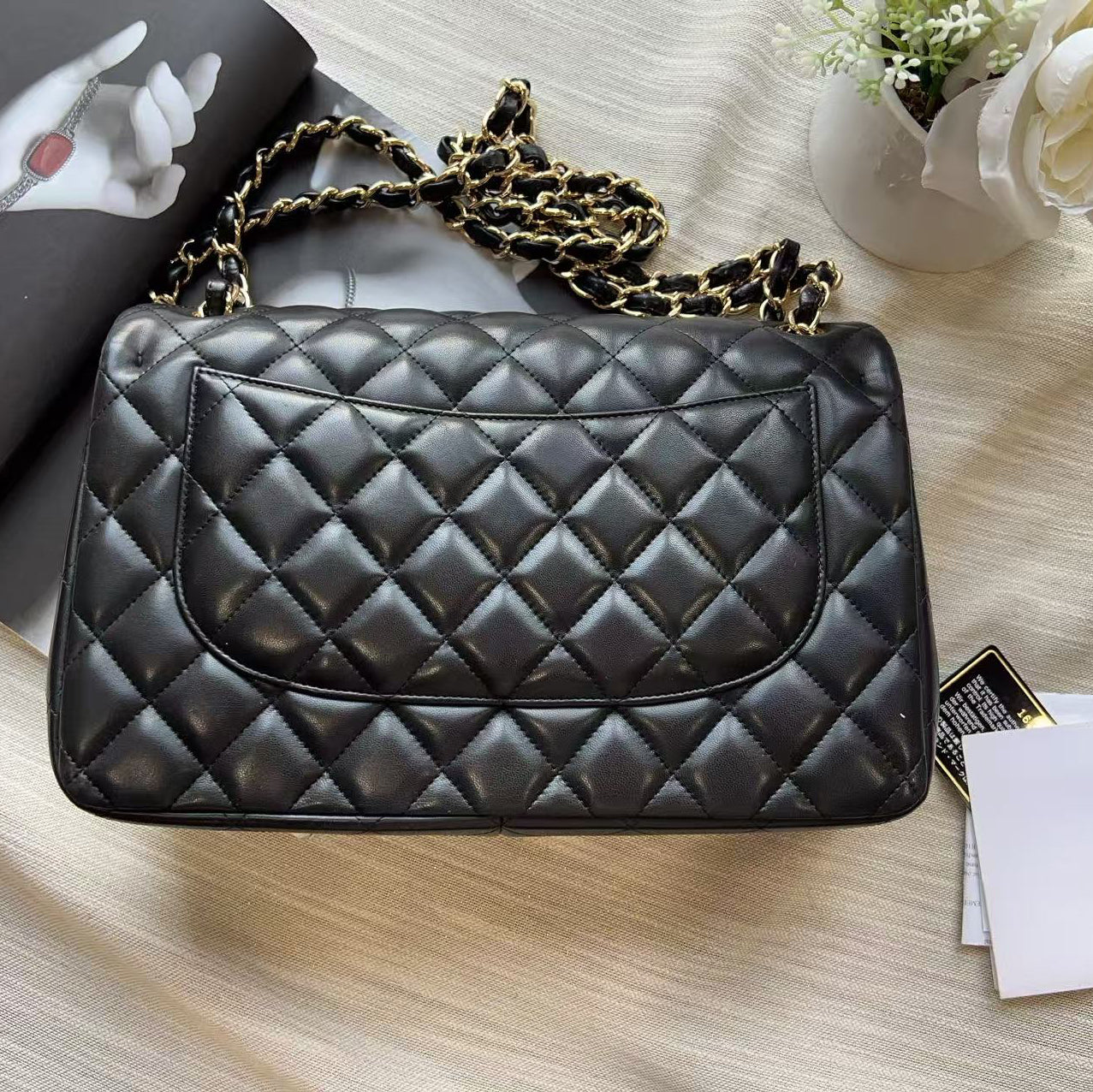 A black quilted leather handbag with a gold chain strap, styled on a beige fabric surface with a magazine, cards, and a white flower.