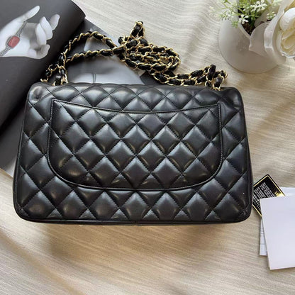 A black quilted leather handbag with a gold chain strap, styled on a beige fabric surface with a magazine, cards, and a white flower.