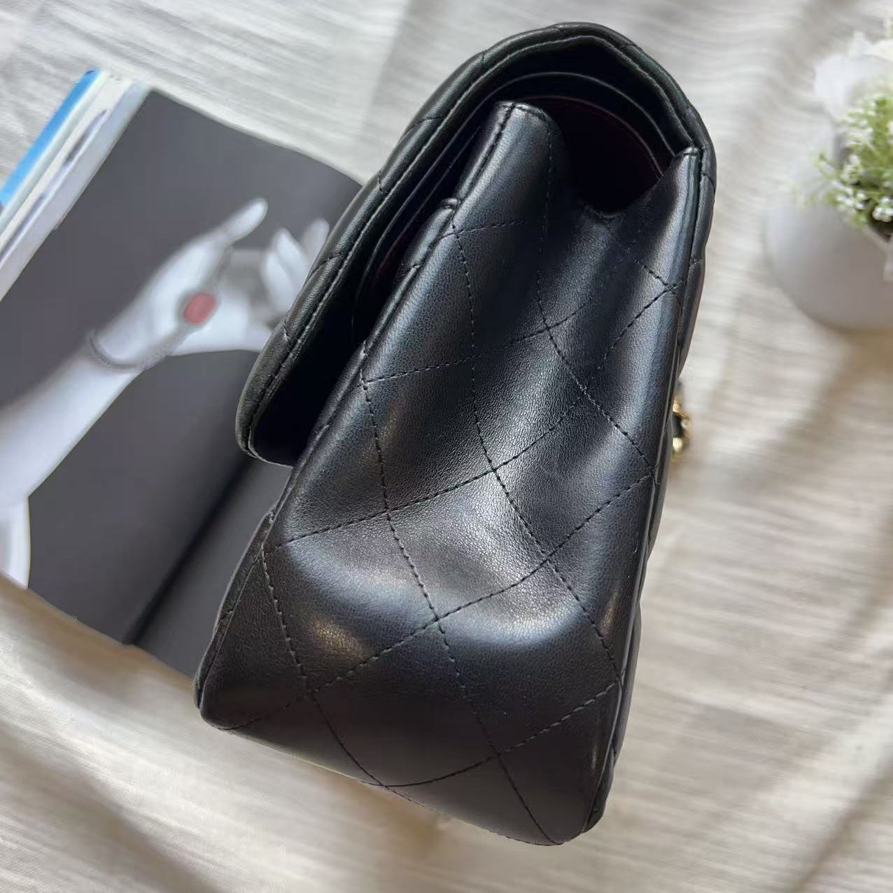 A black quilted leather handbag with a diamond-stitched pattern, photographed from an overhead angle on a textured white fabric surface next to an open magazine.
