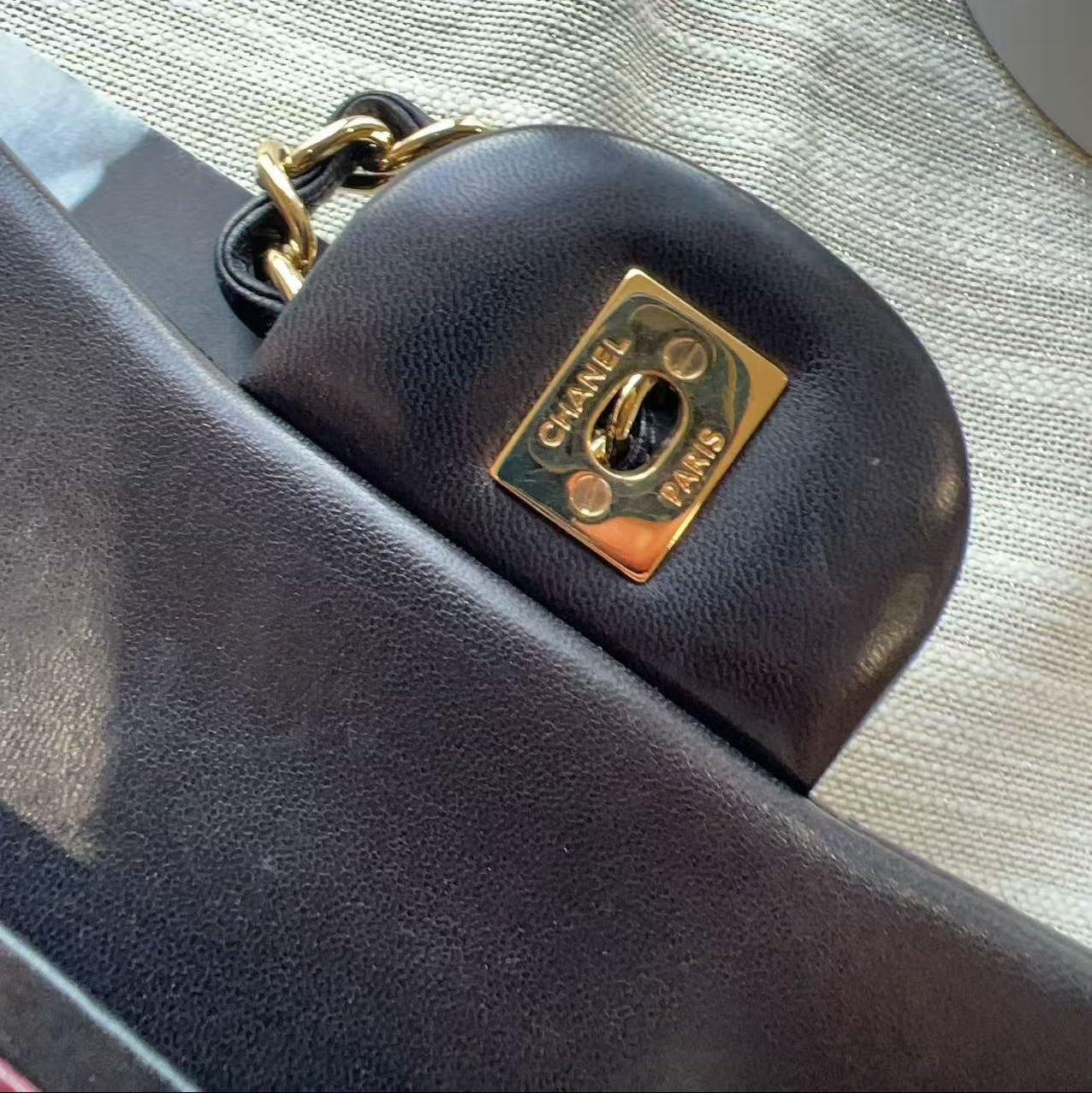 Close-up of a dark brown leather Chanel handbag showing the signature gold-tone CC turn-lock clasp with 'CHANEL PARIS' engraved on a textured beige fabric background.