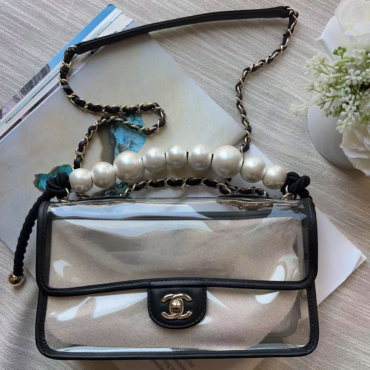 A clear flap shoulder bag with black trim and a gold-tone interlocking CC clasp, styled on top of books with a pearl beaded chain strap and a blue flower charm.