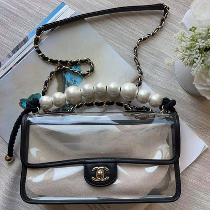 A clear flap shoulder bag with black trim and a gold-tone interlocking CC clasp, styled on top of books with a pearl beaded chain strap and a blue flower charm.