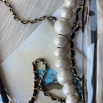 A close-up of a luxury chain and pearl accessory, featuring large white pearls threaded with gold and black chain links, resting on a textured beige fabric surface.