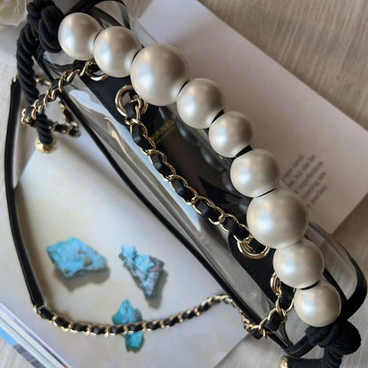 A pair of eyeglasses with a decorative strap featuring large white faux pearls, a black braided cord, and gold-tone chain links, styled on top of open books.