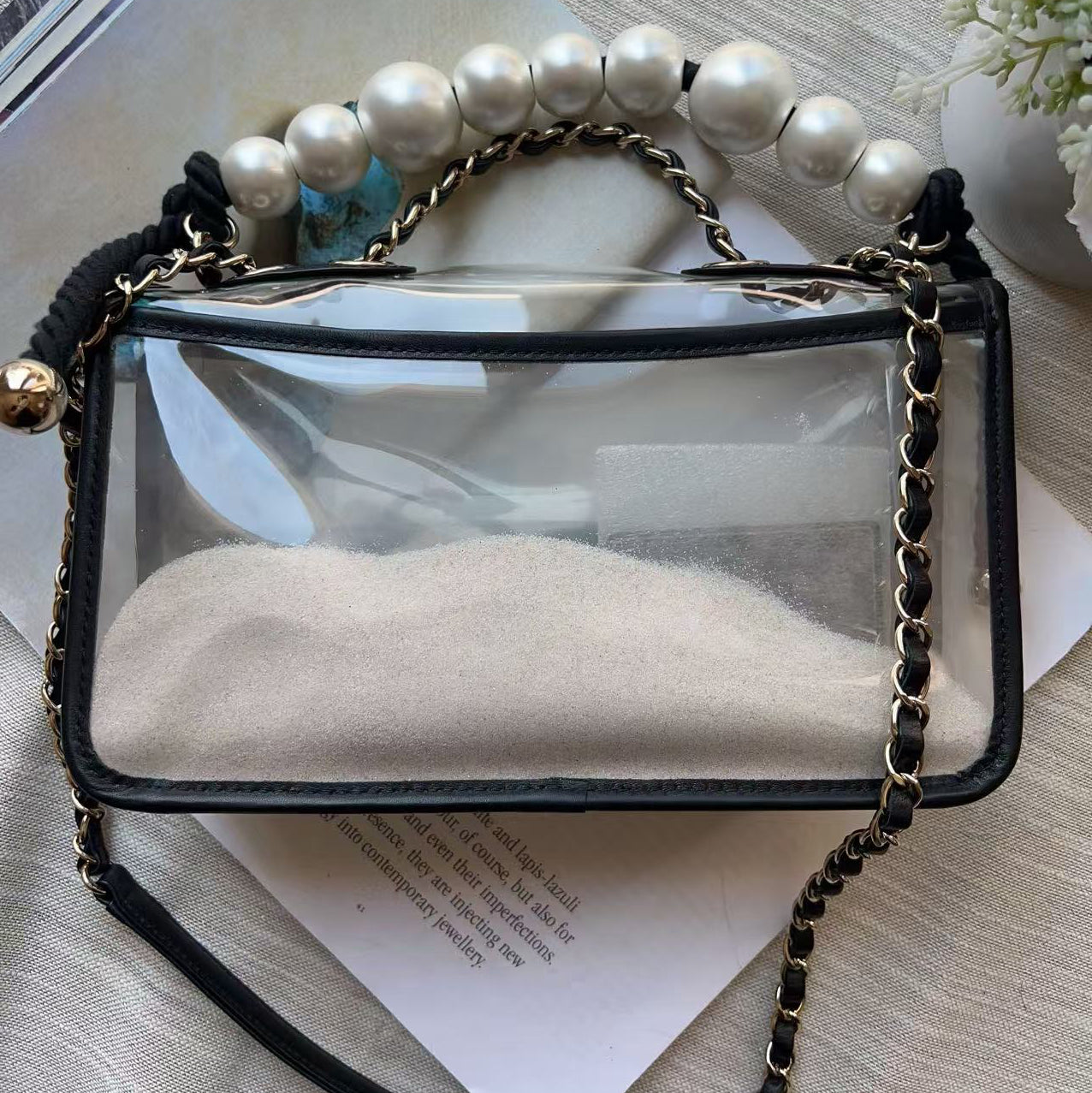 A clear rectangular handbag with black trim and a gold-tone chain strap, styled on top of books with a white pearl bead handle and a small gold bell.