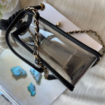 A clear acrylic handbag with black leather trim and a gold chain strap, styled on top of a magazine over a light beige fabric background.