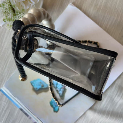 A clear plastic handbag with black trim and a black braided strap, photographed from above on a light fabric surface with a white book and decorative flowers in the background.