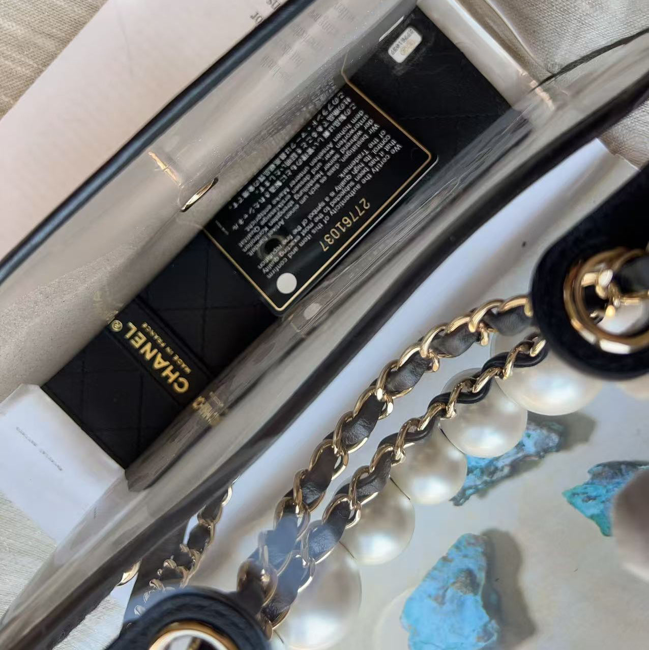 Close-up of a Chanel handbag interior showing a black quilted Chanel card, a clear transparent body, a gold chain strap, and white pearl details.
