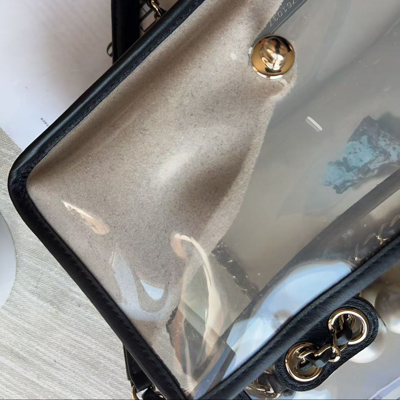 A close-up of a clear vinyl pouch bag with a black border, gold snap button, and black-and-gold chain strap, resting on a light wood surface next to a white book.