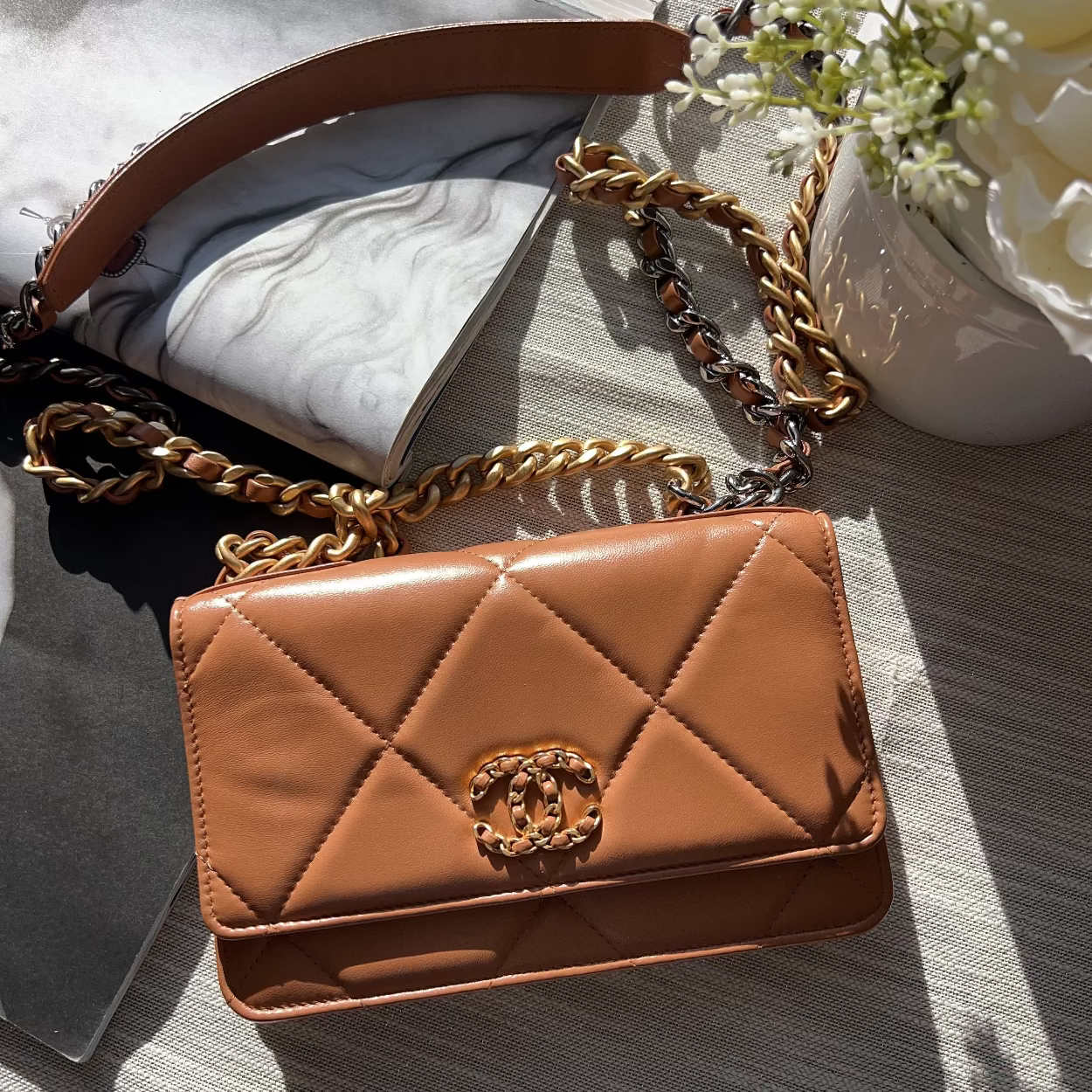 Brown quilted handbag with gold chain strap on a textured surface with flowers.