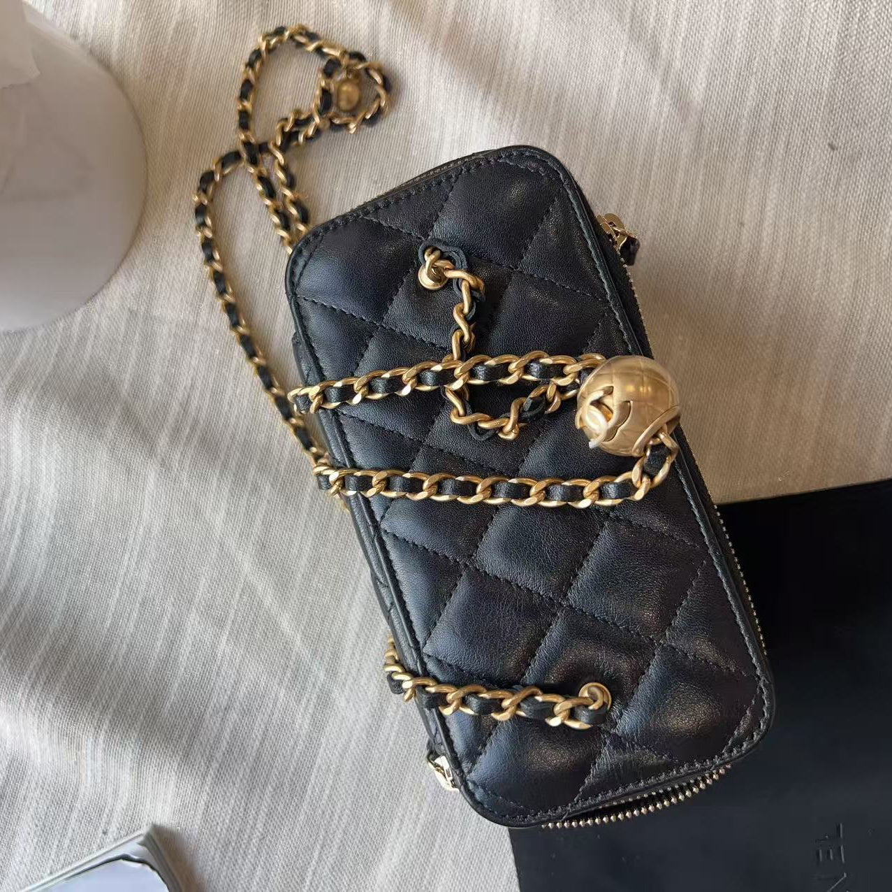 A black quilted leather crossbody bag with gold-tone chain straps and a gold skull charm, laid flat on a textured beige fabric surface.