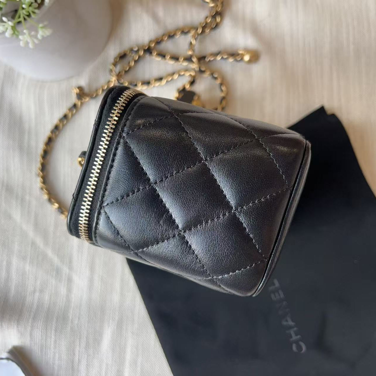 A black quilted leather Chanel crossbody bag with gold-tone chain strap, displayed on a beige fabric surface next to a black Chanel dust bag.