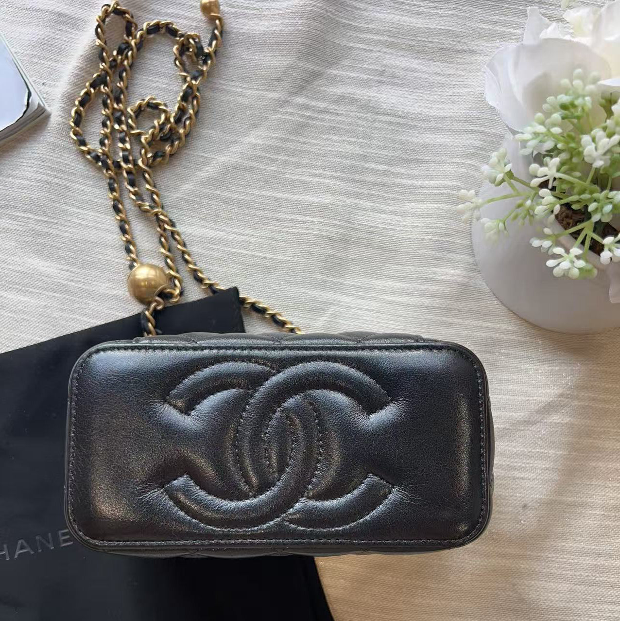 A black leather Chanel phone pouch with an embossed interlocking CC logo, resting on a black Chanel dust bag with a gold chain strap on a textured beige fabric background.
