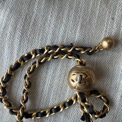 A vintage Chanel shoulder strap featuring a gold-tone chain, black leather inserts, and a central interlocking CC logo charm, laid out on a textured off-white fabric background.