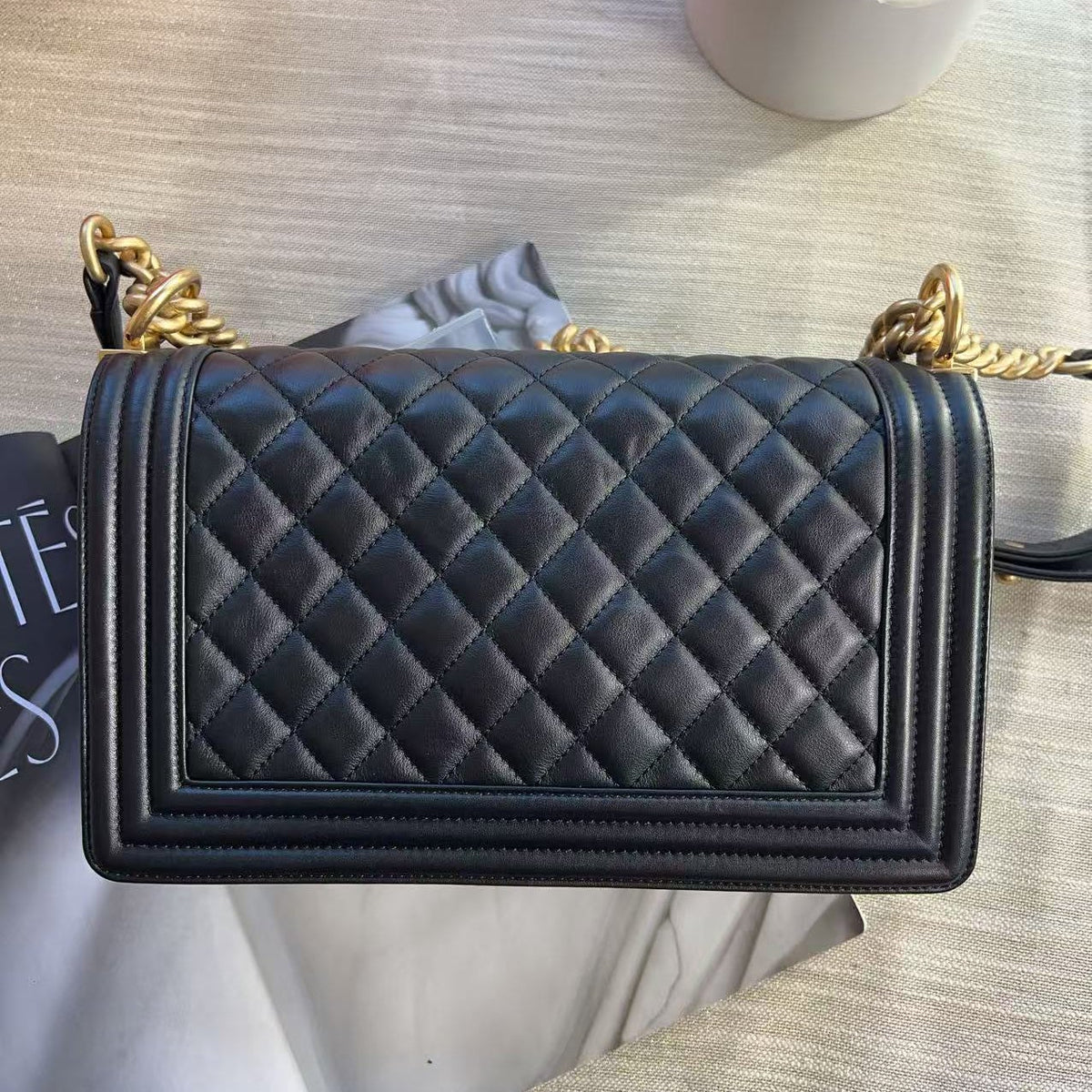 A black quilted leather shoulder bag with gold-tone chain details, shown from a top-down view on a light textured surface with some packaging and decorative props.