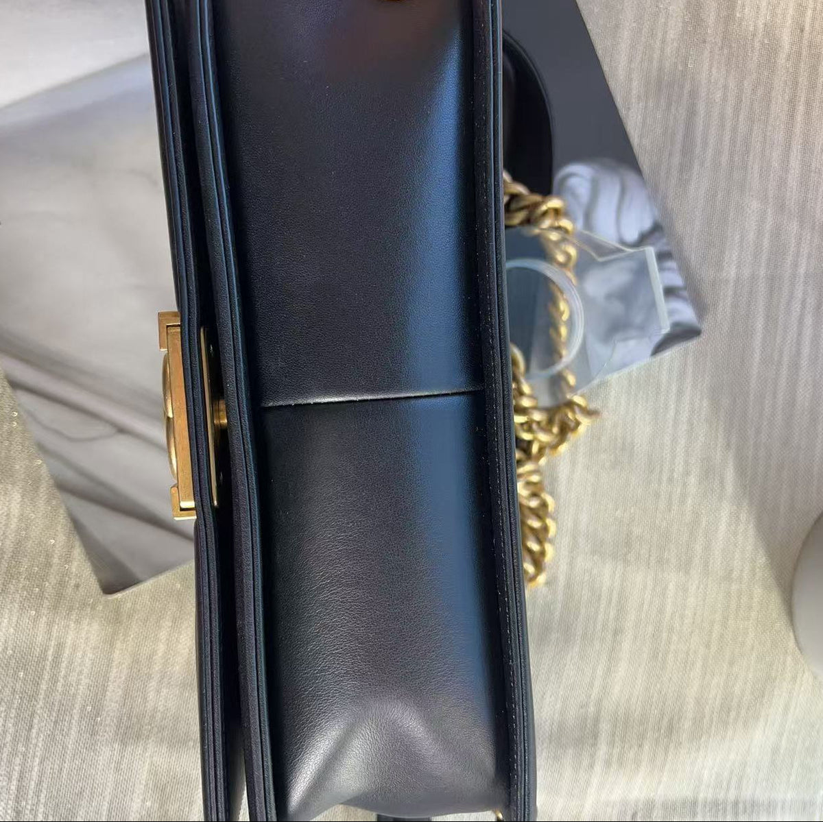 A close-up of a black leather handbag with a gold-tone clasp and a gold chain strap, shown against a light gray textured background.