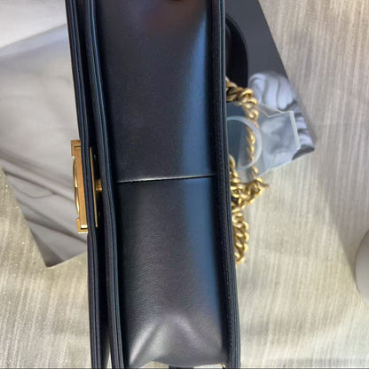A close-up of a black leather handbag with a gold-tone clasp and a gold chain strap, shown against a light gray textured background.