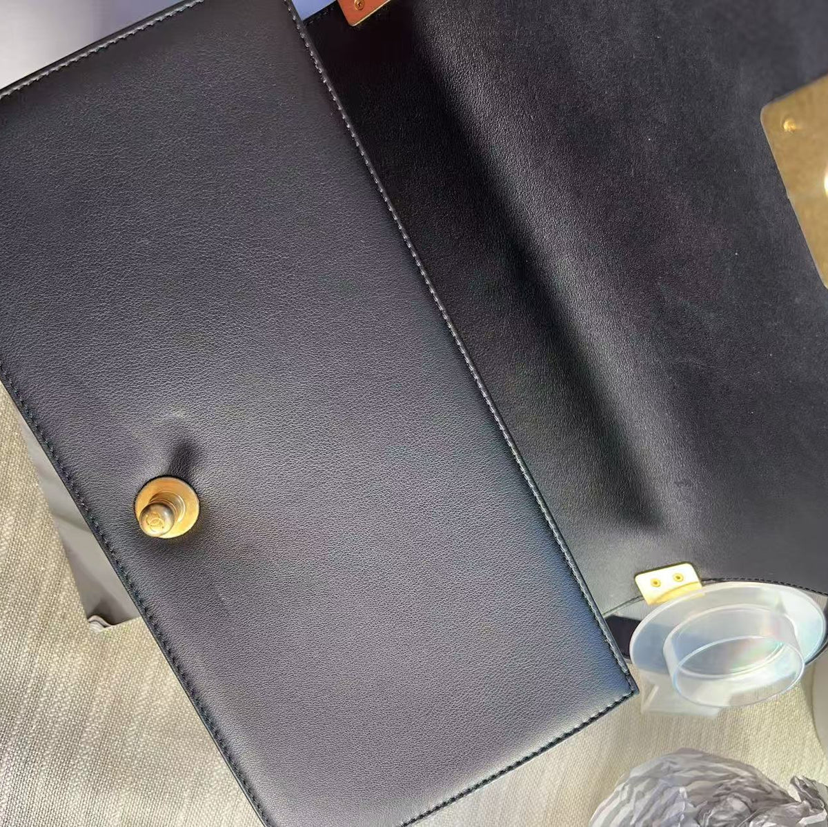 A close-up top-down view of a black leather clutch or wallet with gold-tone hardware, showing the interior flap, snap button, and a small clear plastic insert.