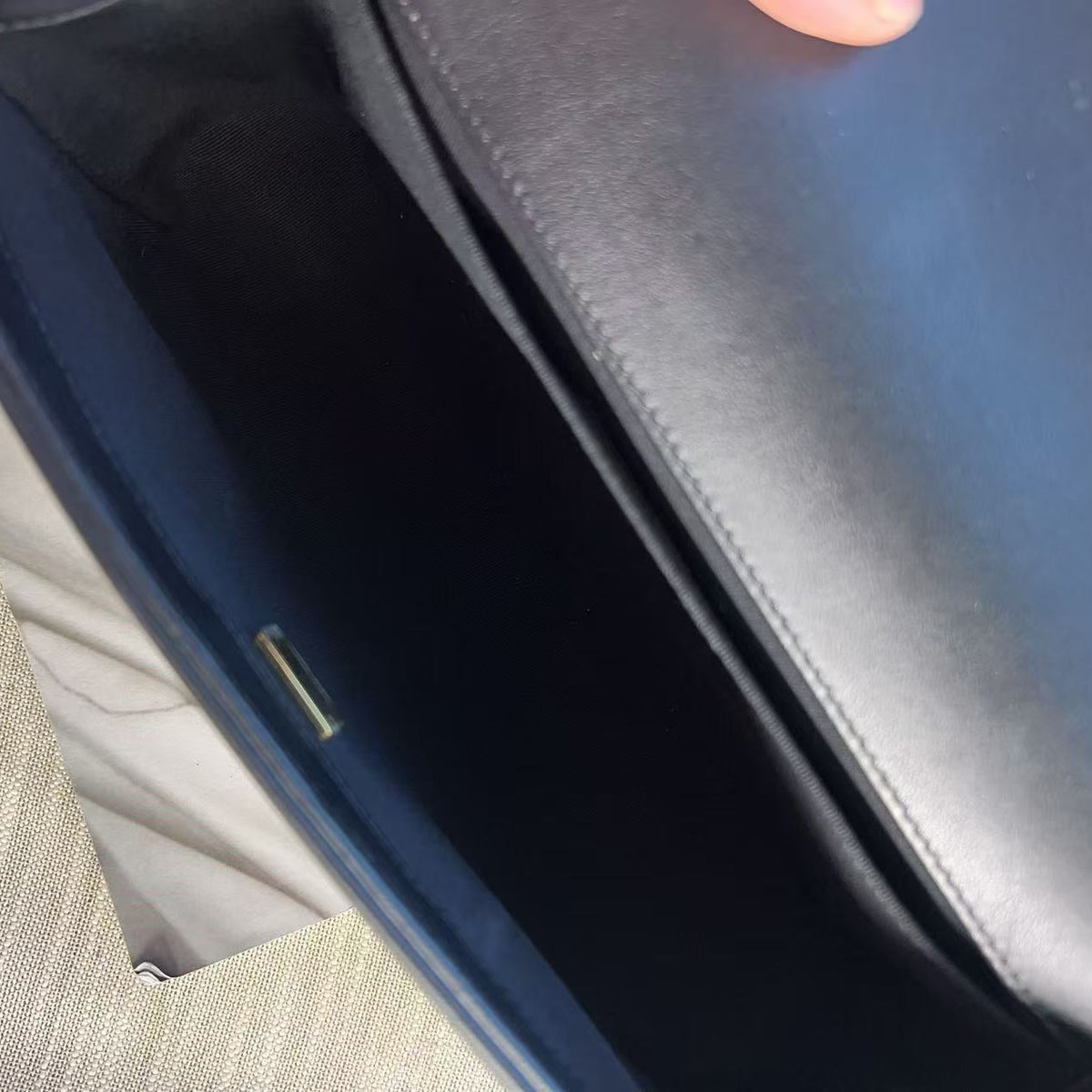 Close-up of a dark navy blue leather bag being opened, with a hand holding the flap and a beige textured fabric surface visible at the side.