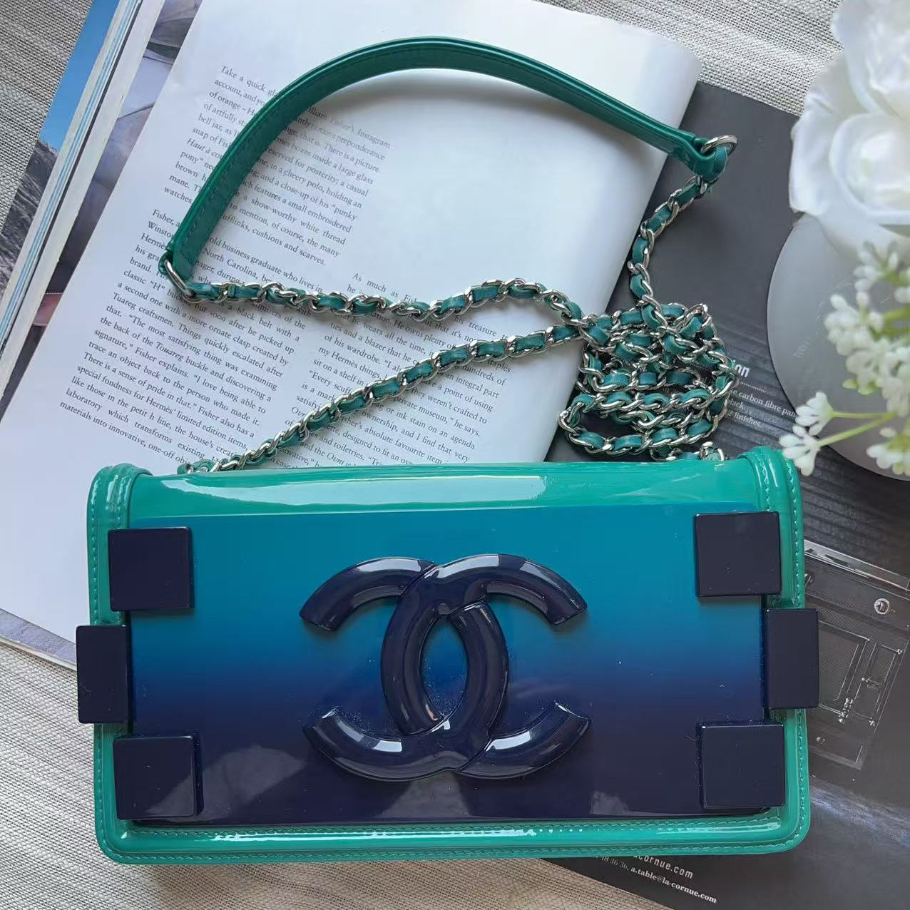A Chanel shoulder bag with a glossy ombre green-to-blue body, featuring the signature interlocking CC logo on the front flap and a green-and-silver chain strap, styled on a light wood surface with an open book and white flowers.
