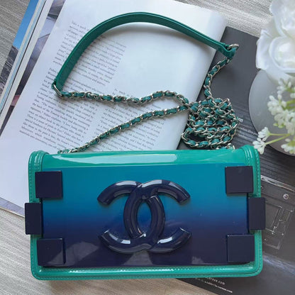 A Chanel shoulder bag with a glossy ombre green-to-blue body, featuring the signature interlocking CC logo on the front flap and a green-and-silver chain strap, styled on a light wood surface with an open book and white flowers.