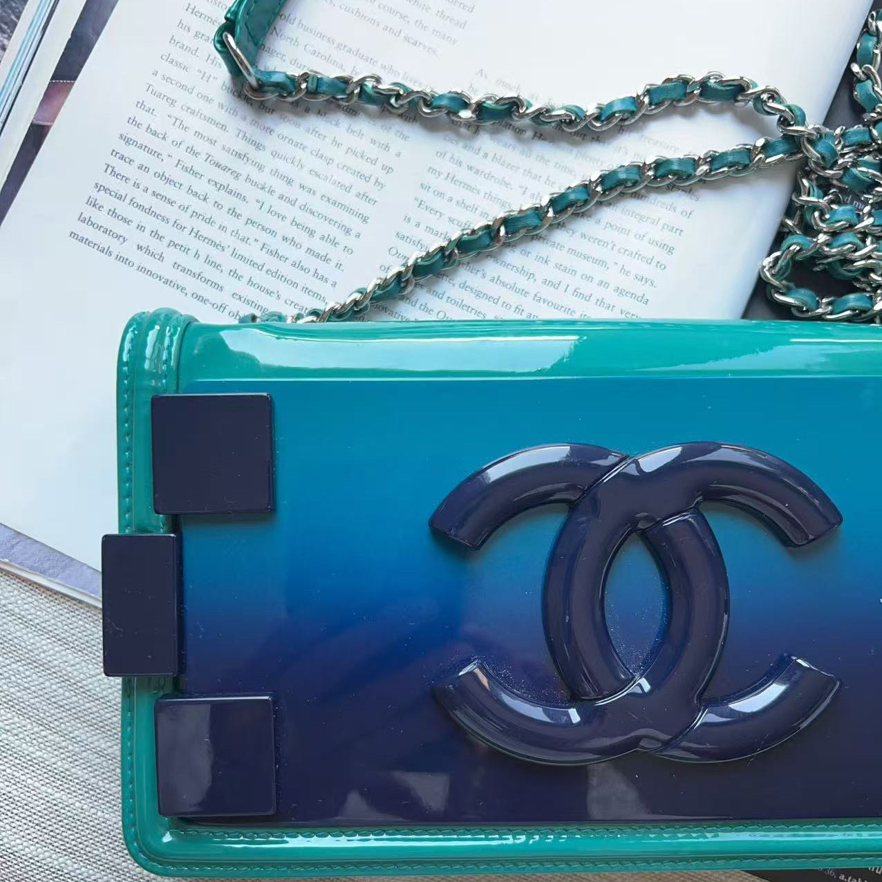 A Chanel shoulder bag with a glossy teal-to-blue gradient patent leather body, featuring a large navy blue interlocking CC logo and a matching chain strap, styled on a textured surface with an open book in the background.
