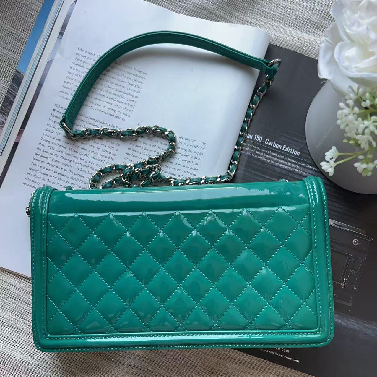 A green quilted patent leather shoulder bag with a chain strap, styled on top of an open book with a white flower vase in the background.