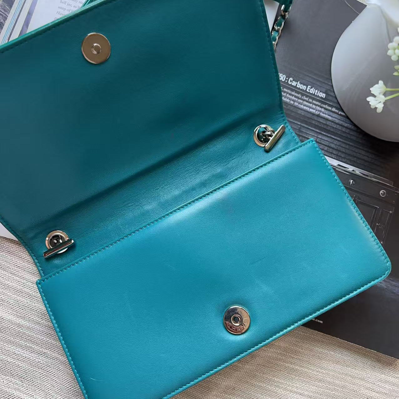 A teal leather clutch handbag shown open, displaying its interior with gold-tone hardware and snap buttons, resting on a light wood surface next to books and a white vase.