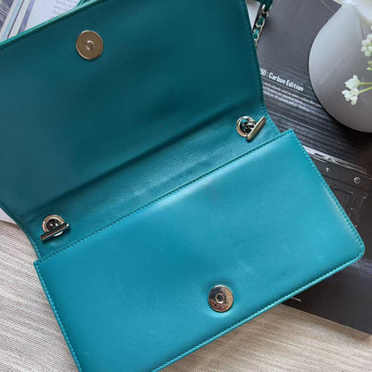 A teal leather clutch handbag shown open, displaying its interior with gold-tone hardware and snap buttons, resting on a light wood surface next to books and a white vase.