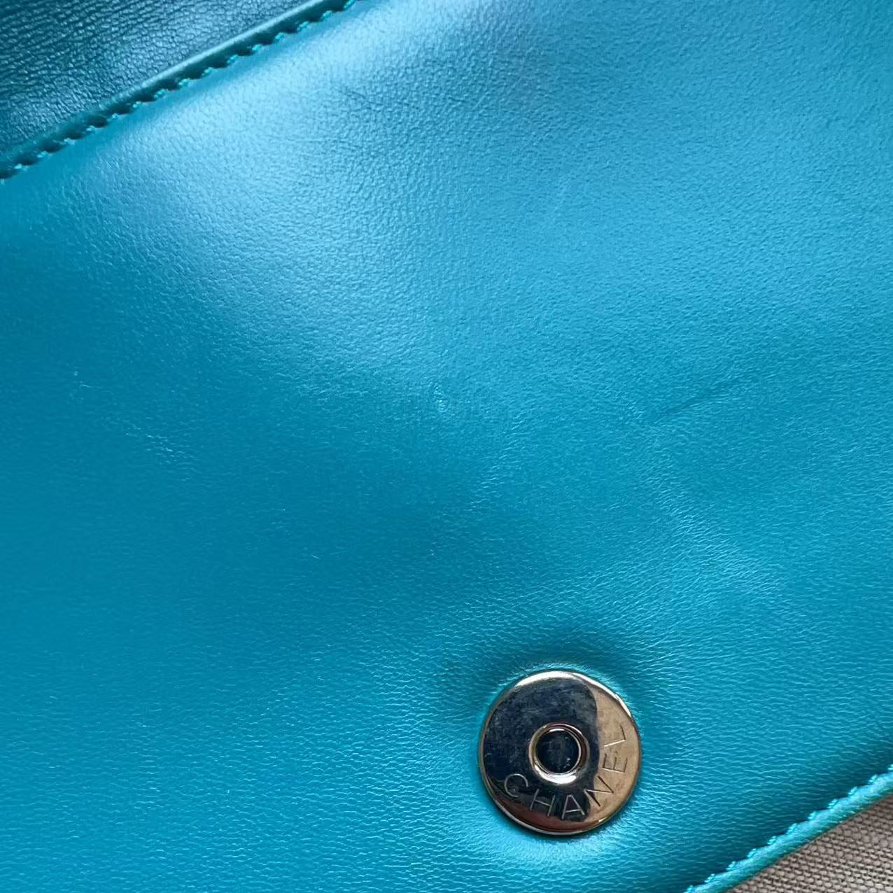 Close-up of a turquoise blue leather clutch or wallet with a silver-tone snap button closure, showing the textured leather surface and stitching.