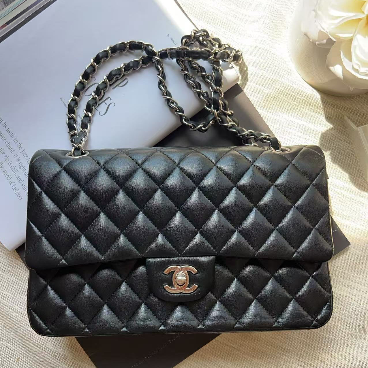 A black quilted leather shoulder bag with a gold-tone interlocking CC logo and a silver-tone chain strap, styled on a light beige fabric surface with books and a white flower.