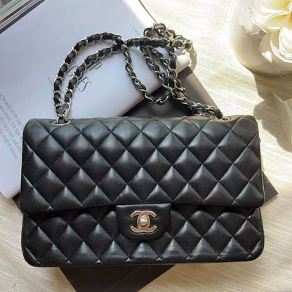 A black quilted leather shoulder bag with a gold-tone interlocking CC logo and a silver-tone chain strap, styled on a light beige fabric surface with books and a white flower.