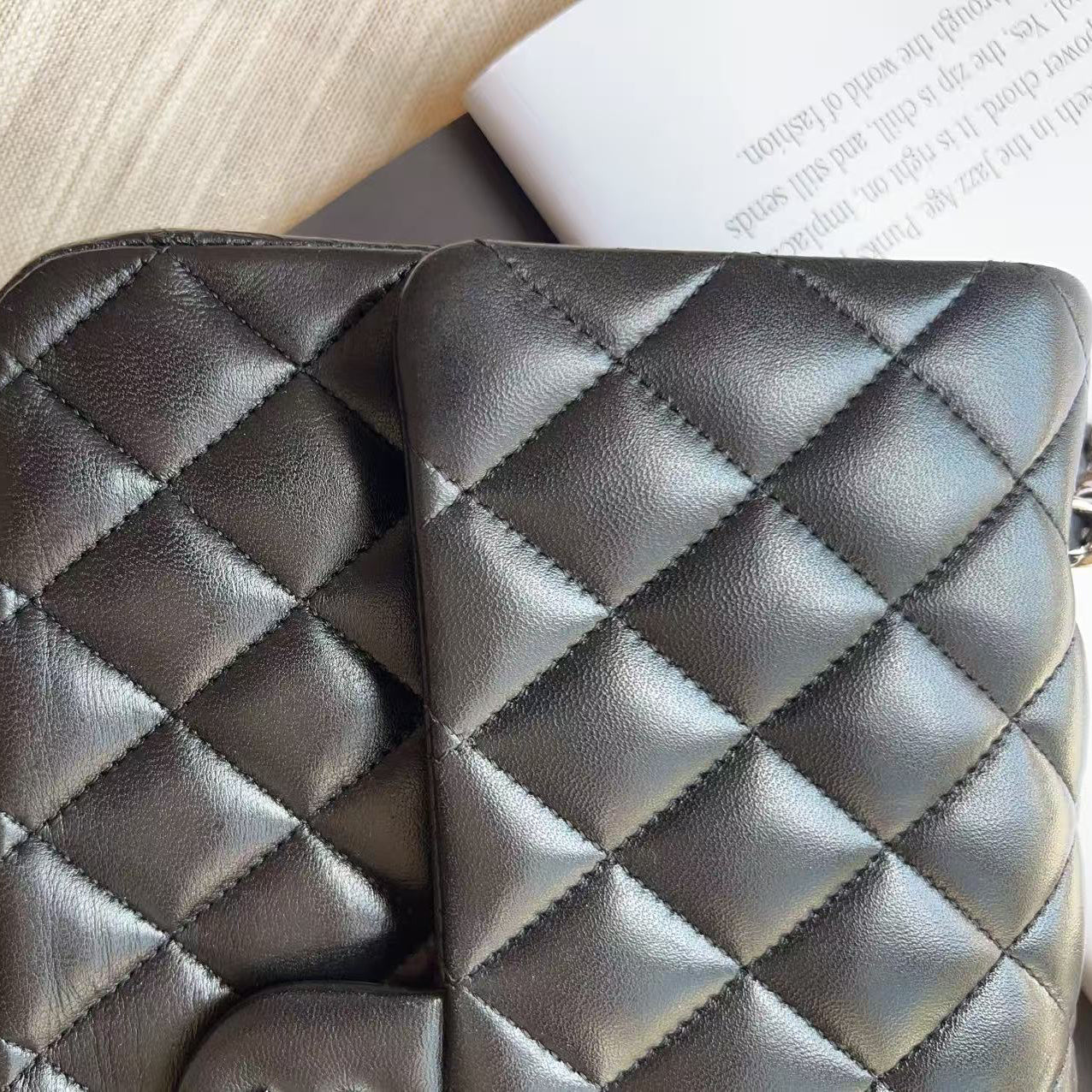 A close-up of a black quilted leather wallet or clutch with diamond stitching, resting on a dark surface next to a book and a light fabric background.