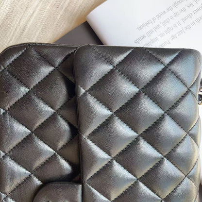 A close-up of a black quilted leather wallet or clutch with diamond stitching, resting on a dark surface next to a book and a light fabric background.