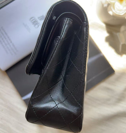 A black quilted leather clutch bag with diamond-stitched detailing, shown from an overhead angle resting on a black box over a beige fabric surface.
