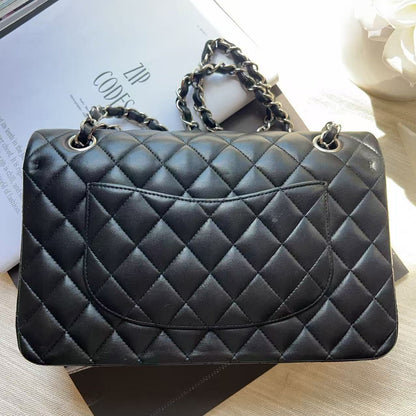 A black quilted leather shoulder bag with silver-tone chain handles, resting on a light beige fabric surface next to a white book or magazine.