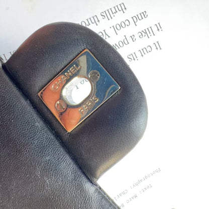 A close-up of a black leather Chanel wallet or clutch featuring the signature square Chanel Paris logo in polished silver-tone metal with a white center, resting on a white printed surface.
