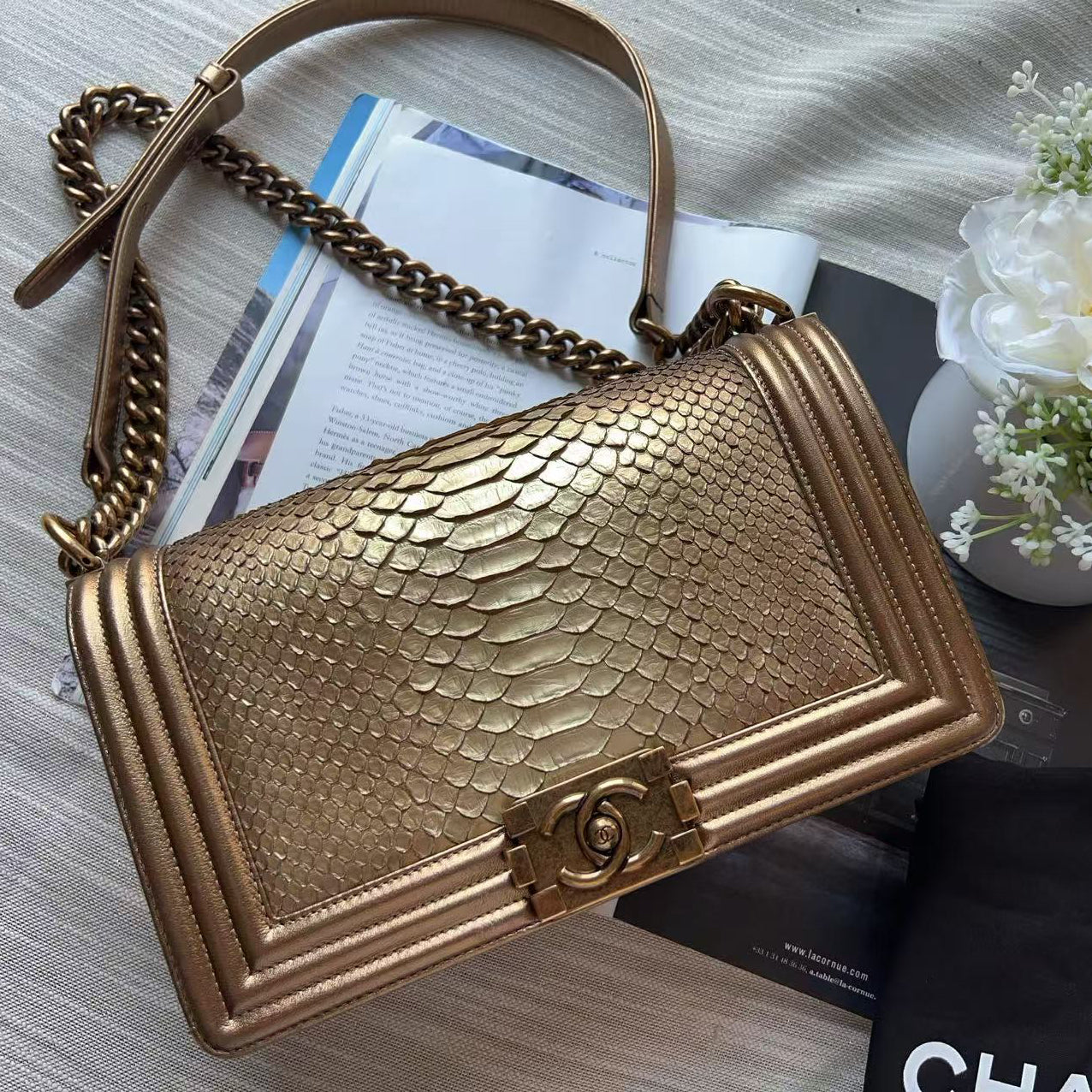 A gold-toned snakeskin-textured shoulder bag with a chain strap, styled on a light fabric surface with a black dust bag, white flowers, and printed papers.