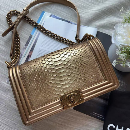 A gold-toned snakeskin-textured shoulder bag with a chain strap, styled on a light fabric surface with a black dust bag, white flowers, and printed papers.