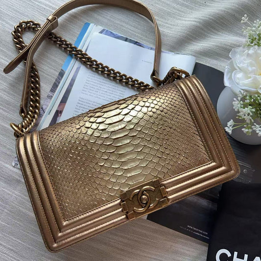 A gold-toned snakeskin-textured shoulder bag with a chain strap, styled on a light fabric surface with a black dust bag, white flowers, and printed papers.