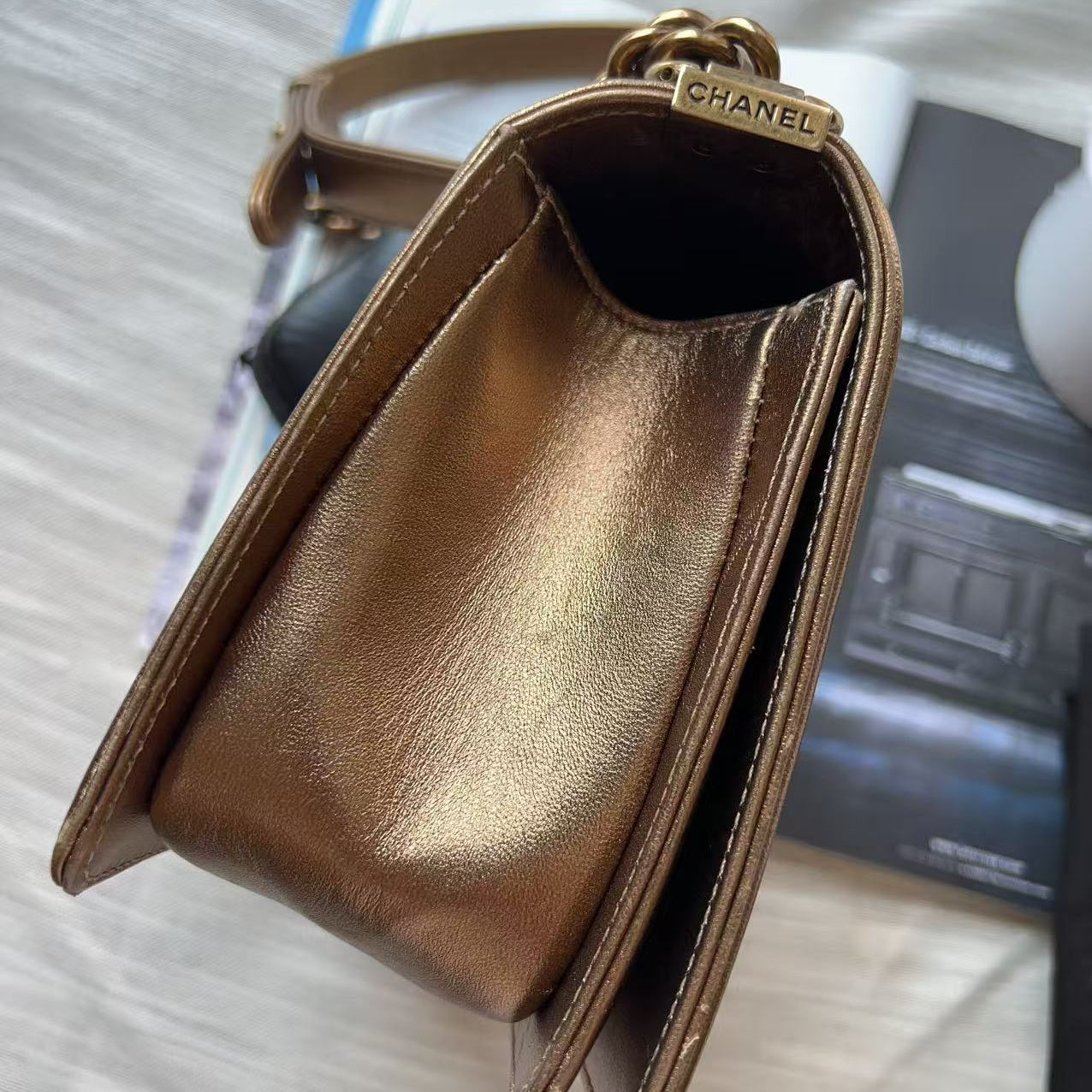 A Chanel shoulder bag in metallic bronze leather, shown from an overhead angle resting on a light-colored fabric surface next to an open magazine. The gold-tone metal clasp features the CHANEL logo.
