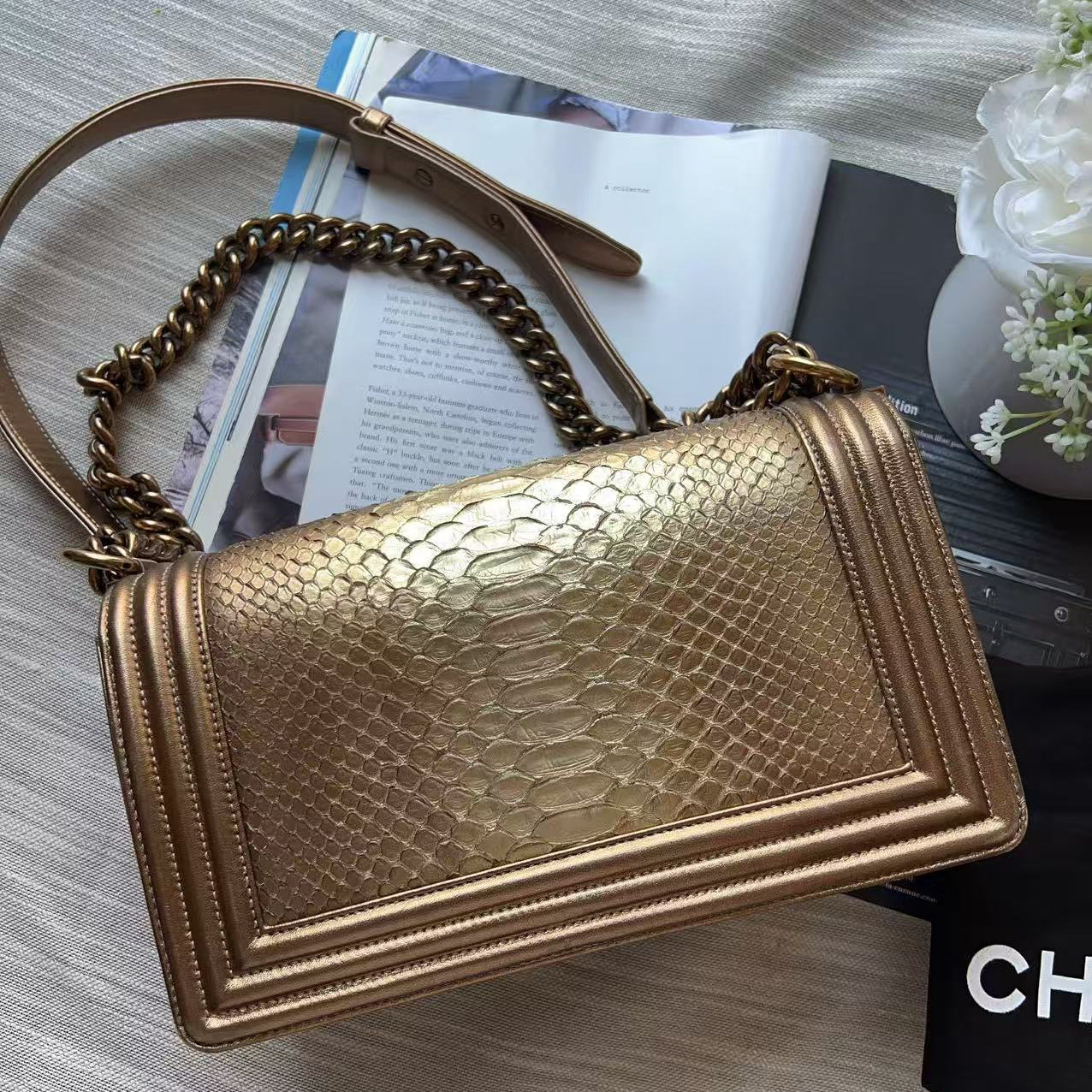 A metallic gold shoulder bag with a textured python-style embossed pattern, featuring a gold chain strap and a gold-toned top handle, styled on a light gray fabric surface with a magazine, black box, and white flowers.